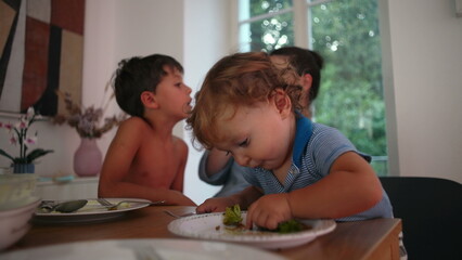 Fototapeta premium Toddler eating broccoli while sitting at dining table next to older brother during family meal, concept of healthy nutrition, childhood development and sibling togetherness