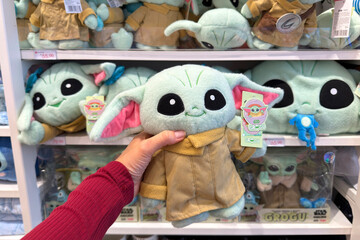 Naklejka premium Dubai, UAE - 24 may 2026: Hand holding Grogu plush character in modern toy store surrounded by collectible Star Wars merchandise, representing entertainment retail and fandom culture lifestyle