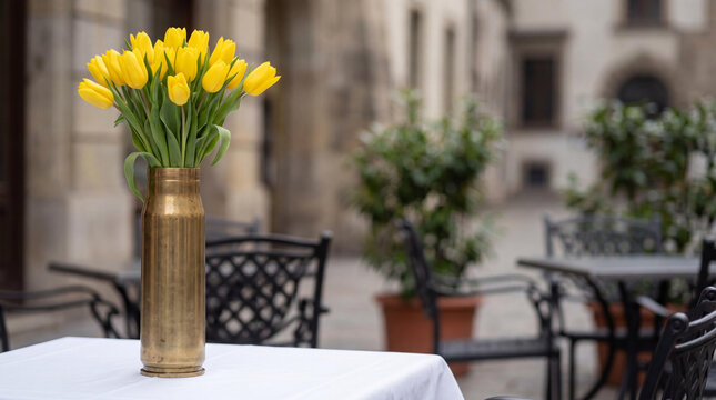 Yellow tulips in brass shell casing as vase. Symbol of peace after war. Flower arrangement on table in street cafe. Hope, rebirth and resilience in post conflict city scenery.