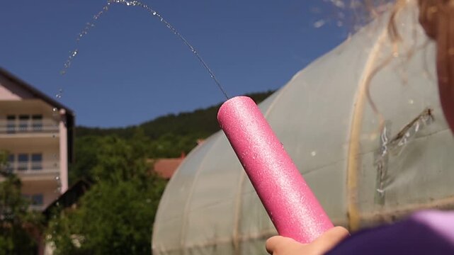 Child sprays a stream of water from a colorful foam water blaster outdoors on a sunny day, creating a playful arc of droplets in a backyard near a greenhouse and residential home.