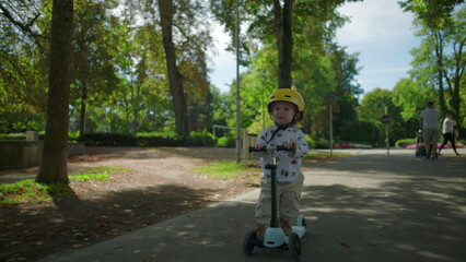 Obraz premium Little child with yellow helmet riding scooter along tree-lined path in park, moving forward with curiosity and balance in natural outdoor environment