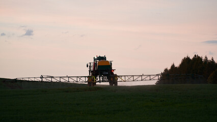 Fototapeta premium A self-propelled sprayer sprays pesticide, herbicide or liquid mineral fertilizer onto green shoots in the field in the evening at sunset.