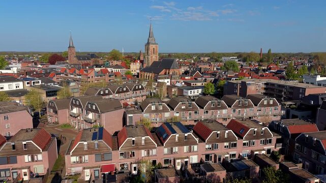 An aerial drone view of a European town landscape showcasing rows of modern brick houses with distinct mansard roofs, looking toward historic church spires rising above the city center.