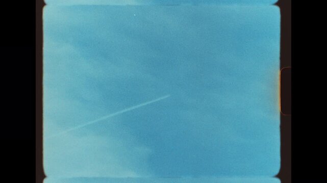 A small high altitude jet aircraft flying from left to right across a vivid blue sky leaving a faint white vapor trail, filmed on retro super 8 film