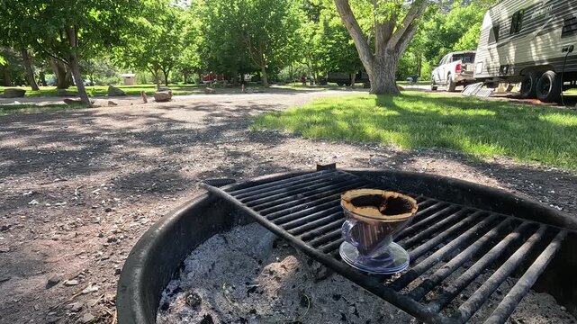 A fresh coffee dripper sitting on a campfire grill at a scenic campground.