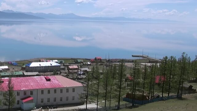 Breathtaking vista of a serene Mongolian village hugging the vibrant shores of Lake Khuvsgul. Cloud patterns and distant mountains dance upon the mirror-like blue waters under a bright sun.