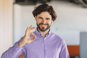 Fototapeta premium A man stands in an office environment. He is smiling and giving an okay sign with his right hand. The setting has warm colors and a modern feel. He appears happy and confident
