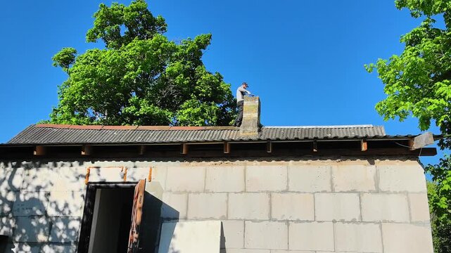 A worker dismantles an old chimney on the roof of a house in the village. A worker is on top of a building roof placing materials on the chimney. The sun shines brightly and trees are visible nearby. 