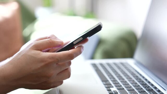 Close up of hand typing on smartphone beside laptop. Mobile messaging, online communication, customer support, digital interaction and modern business technology workflow.
