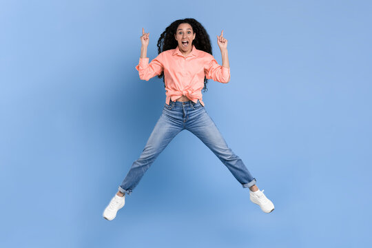 A woman is jumping with joy against a blue wall. She is wearing a pink shirt and jeans. Her hair is curly and she has a big smile on her face, expressing happiness and energy