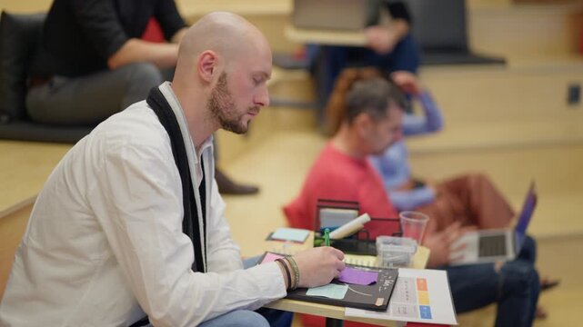 A bald professional takes notes during a focused business meeting. A young employee sits in a modern collaborative environment