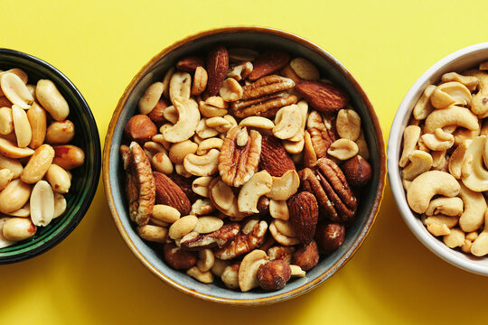 Mixed nuts in bowls on yellow background. Assorted healthy snack selection. Almonds cashews peanuts and pecans. Protein rich food flat lay composition.