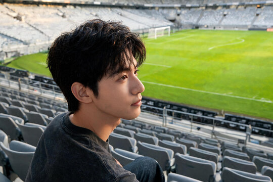 Joven asi&aacute;tico sentado en las gradas de un estadio de f&uacute;tbol vac&iacute;o mirando hacia el campo verde. Retrato de perfil de un aficionado, jugador o espectador en un entorno deportivo moderno.