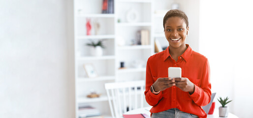 A woman stands in a bright room, holding a smartphone and smiling. The space has bookshelves and plants. Sunlight fills the area, creating a lively atmosphere © Prostock-studio