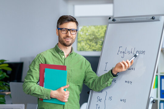 Smiling Caucasian Male Teacher Engaged In Teaching English Lesson With Whiteboard In Modern Office Setting, Holding Materials And Marker, Focused On Educating Students. Education Concept