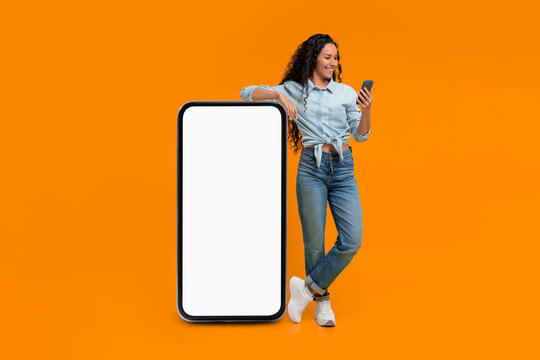 A young woman stands beside a large blank screen while using her smartphone. She smiles as she engages with her device, and the background is bright orange