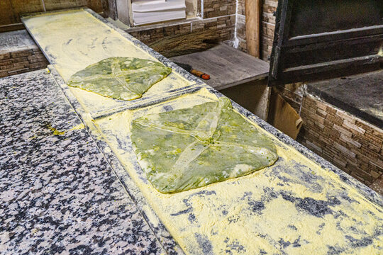 Chef spreading crushed green pistachios and fresh clotted cream on thin dough to prepare traditional Antep Katmeri pastry, close up view.