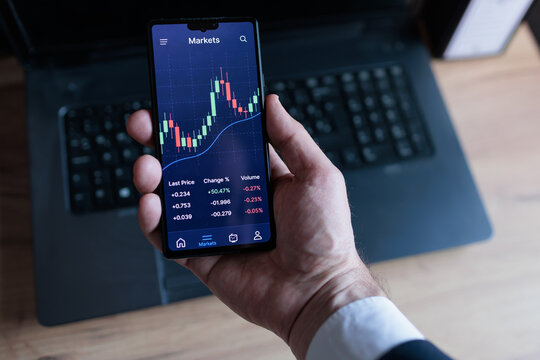 Businessman uses smartphone to check stock market data with a laptop in background