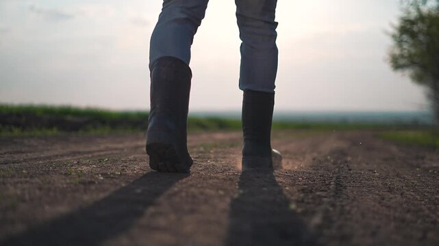 Farmer boots press into dirt field path while walking. Calm outdoor environment frames walking motion. Black boots lift and lower on dry path. Legs show walking balance of farmer on dirt ground.