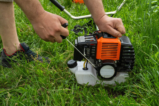 A gardener pulls the starting cord of a lawn mower in a grassy yard. The sun shines in the background, and hands grasp the equipment tightly. The mower is positioned on the ground