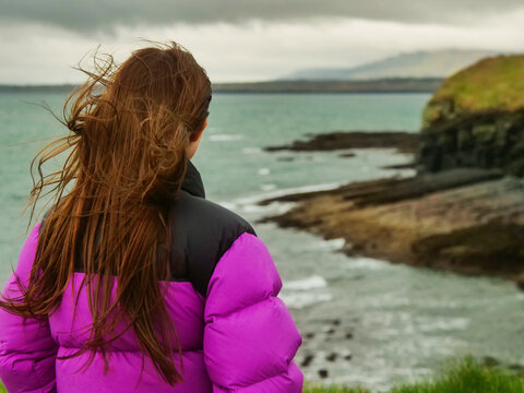 A woman stands on a grassy hill overlooking the ocean and cliffs. She is wearing a purple jacket and black pants. Mullaghmore area in county Sligo, Ireland. Travel and tourism. Cloudy sky.
