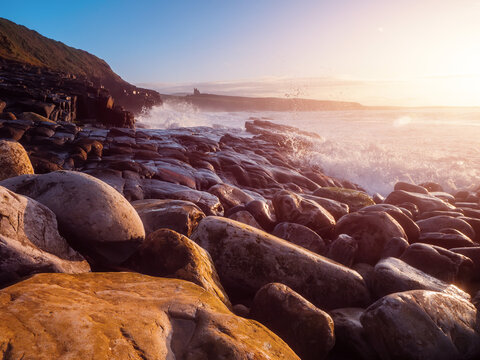 Sunlight glows over foamy ocean waves. Foam blankets pebbled shoreline, soft and golden. Rocks and grassy cliffs frame the scene. Mist hovers near water edge, gentle and serene. County Sligo, Ireland.