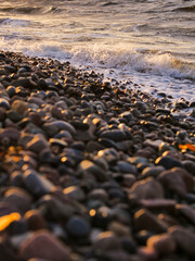 Obraz premium Golden light kisses smooth pebbles at water's edge. Waves crash gently, leaving foam trails behind. Rocks gleam with warm hues, reflecting sun's glow. Rough ocean coast.
