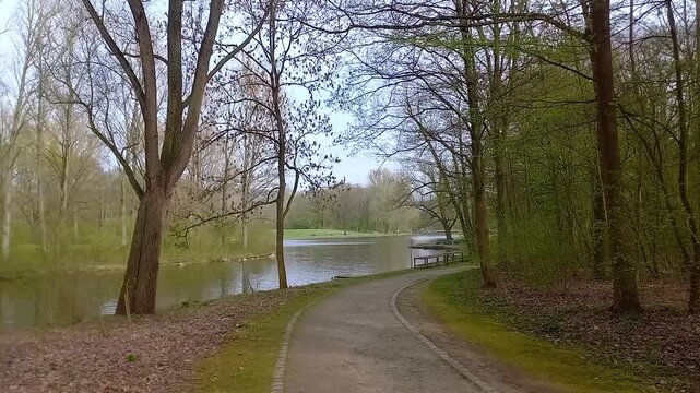 First-person view (POV) of holding a camera and walking down a winding alley path towards a calm lake surrounded by trees in early spring at Freizeitpark Rheinaue in Bonn, Germany.