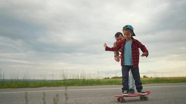 Father guiding son on skateboard outdoors: Father helps son maintain balance on skateboard. Boy enjoys outdoor skateboarding with father guidance. Family learning skateboarding together outside.