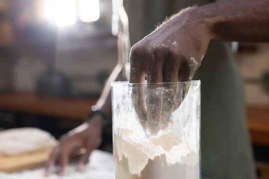 African American adult man using right hand in clear flour jar, shaping dough on wooden board