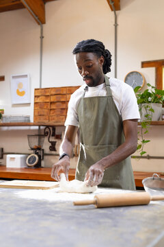 African American man wearing green apron shaping dough on floured bakery counter with rolling pin