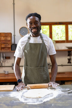 African American man in olive apron smiling, rolling dough with rolling pin in bakery workspace