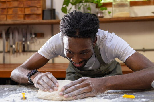 Adult African man wearing olive apron kneading dough on floured wooden counter, yellow clip