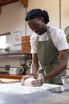 Man kneading dough at bakery kitchen wearing olive-green apron, wooden board left, metal bowl right