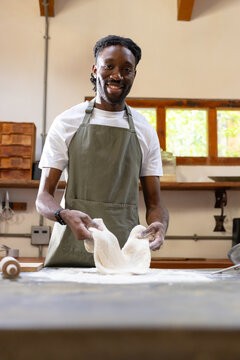 Adult male baker wearing olive apron stretching dough, smiling on bench with rolling pin at bakery