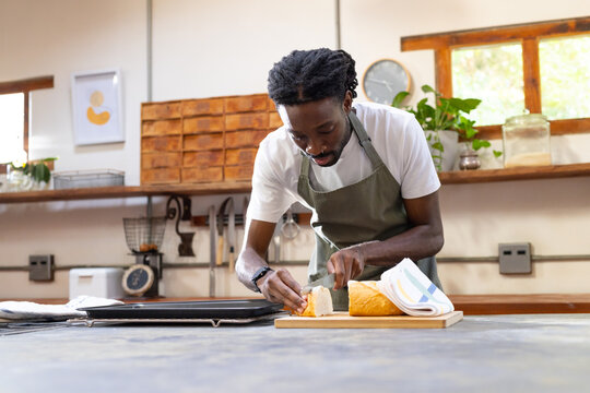 African American man in green apron slicing loaf of bread on wooden counter with knife
