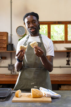 African American man wearing green apron at wooden counter in small bakery, holding torn loaf