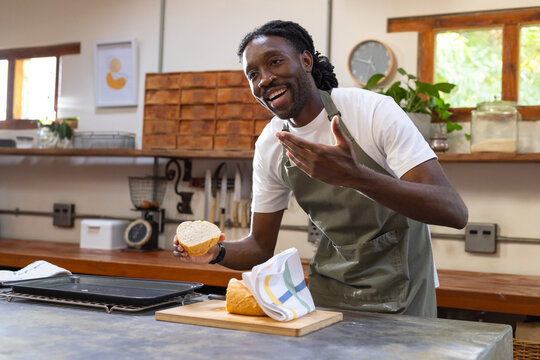 Adult African man wearing green apron holding half loaf, gesturing in home kitchen by cutting board