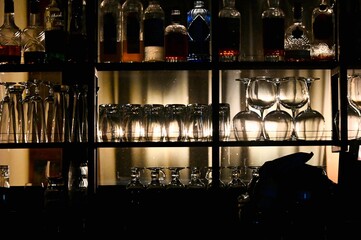 A beautiful horizontal shot of a bar counter shelf illuminated by warm backlighting, displaying rows of clean, upside-down wine glasses, cocktail glassware, and various liquor bottles © Drachenfutter