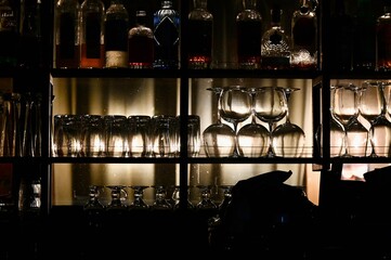 A beautiful horizontal shot of a bar counter shelf illuminated by warm backlighting, displaying rows of clean, upside-down wine glasses, cocktail glassware, and various liquor bottles © Drachenfutter