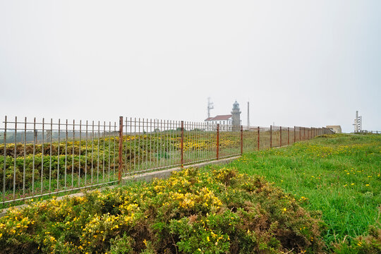Cabo de Penas lighthouse and communication buildings behind a metal fence, set among green grass and yellow gorse under a misty coastal sky, travel and nature scene