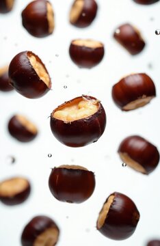 Brown horse chestnuts float in air against white background. Some nuts split open revealing light inner part. Tiny water droplets visible around.