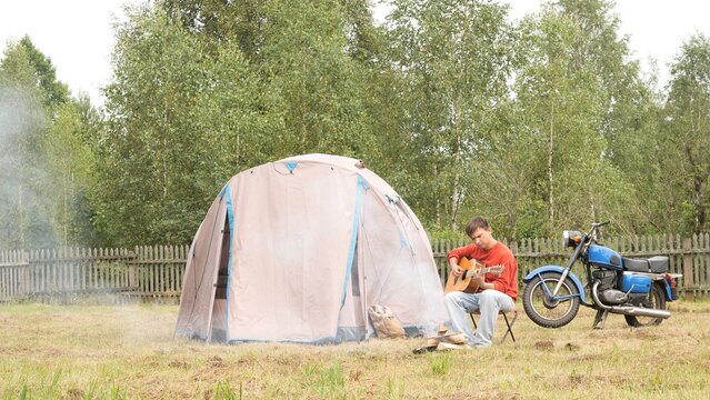 Young man beside his tent and motorcycle, sitting on grass playing acoustic guitar, enjoying peaceful solo road trip in forest wilderness, relaxed and unplugged from routine