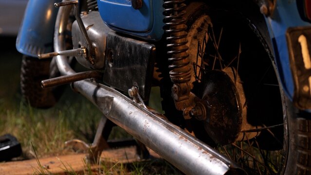 Close up of vintage blue motorcycle rear wheel, rusty exhaust and worn shock absorber at night, chrome and patina details highlighting aged mechanical character and street nostalgia
