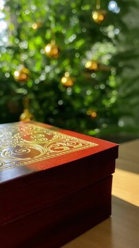 Red wooden box with gold patterns on a table. Vertical video of a festive gift box with a blurred Christmas tree background. Holiday celebration concept