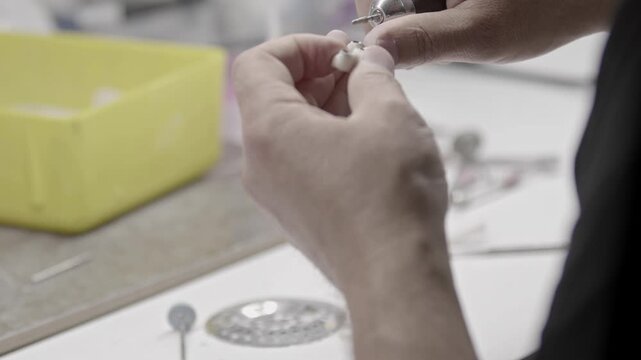 Close up of dental technician hands carefully making precision adjustments to a white zirconia ceramic tooth crown using a special micro handpiece tool in a laboratory.