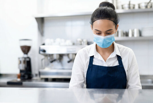 Female cafe worker in an apron and face mask standing in a coffee shop kitchen
