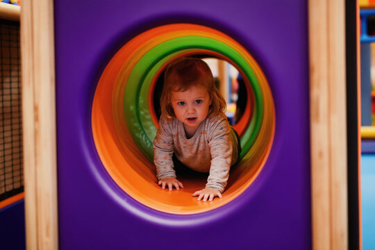 Small girl crawling through colorful indoor playground tunnel. Toddler plays in kids zone with soft toys. Joyful child enjoys fun activity in playroom.