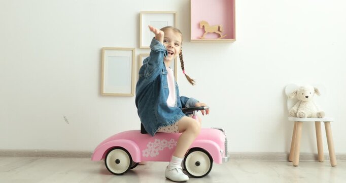 Little girl driving pink toy car near white wall at home