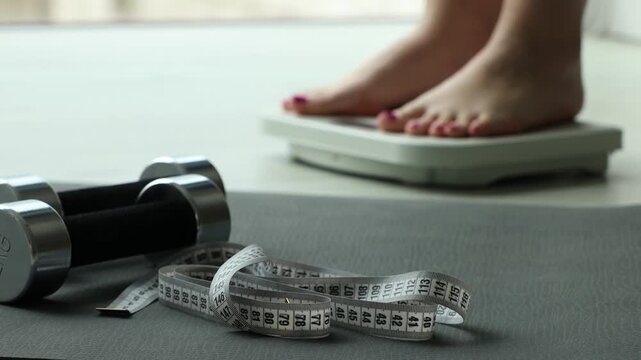 Woman standing on scales indoors, focus on measuring tape, dumbbells and fitness mat